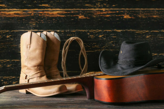 Cowboy boots, hat, and guitar on wooden surface.