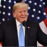 Man speaking at podium with American flags background.
