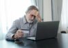 Elderly man looking frustrated while holding a credit card and sitting in front of a laptop