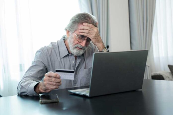 Elderly man looking frustrated while holding a credit card and sitting in front of a laptop