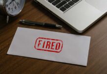 An envelope marked 'FIRED' on a wooden desk next to a clock and a laptop
