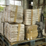 Stacked packages wrapped in plastic next to a rifle in a warehouse setting