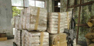 Stacked packages wrapped in plastic next to a rifle in a warehouse setting