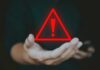 Russian Mafia HIJACKS Hospice System for Billions A hand holding a red warning triangle with an exclamation mark