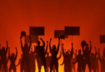 Massive Protest Wave Hits All 50 States Silhouetted crowd of protesters holding signs against a fiery background