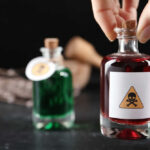 A hand holding a glass bottle labeled with a poison symbol, next to another bottle