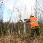 Hunters Exposed — ID Goldmine Unlocked Hunter in high-visibility vest aiming rifle in forest.