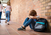 Measles Danger Creeps Into Classrooms? Sad child sitting against brick wall, kids running away.