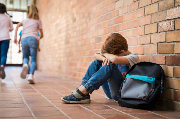 shutterstock_1136889179 Sad child sitting against brick wall, kids running away.