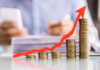 Profit Shock Rockets Wall Street Stacked coins with upward red arrow graph in foreground.