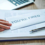 Brutal Cuts Hit Iconic Car Company A hand resting on an envelope labeled 'YOU'RE FIRED' on a desk with papers and a pen