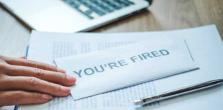A hand resting on an envelope labeled 'YOU'RE FIRED' on a desk with papers and a pen