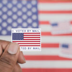 Hegseth Draws A Hard Election Line Hand holding a vote-by-mail sticker with U.S. flag