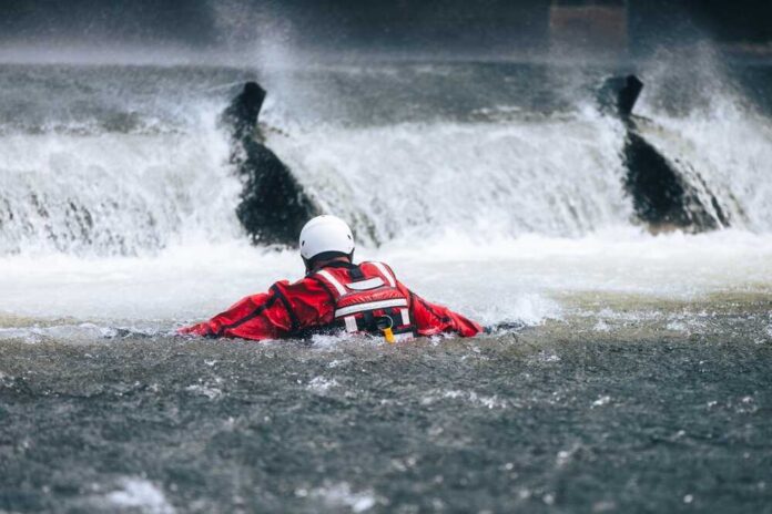 Firefighter,In,Water,Under,Dangerous,Wier,During,Drowning,Rescue,Training. Rescuer in red gear navigating turbulent water near a waterfall