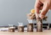 Stacks of coins with shopping cart icons and a hand placing one on the tallest stack