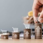 Stacks of coins with shopping cart icons and a hand placing one on the tallest stack